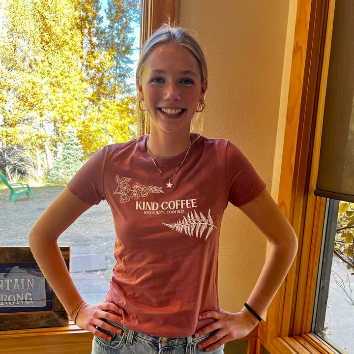 Smiling girl in a t-shirt that has two floral elements and "Kind Coffee Estes Park, Colorado" in the center 