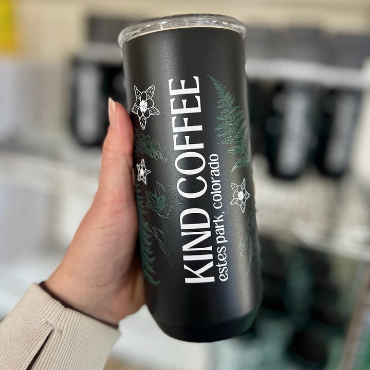 A person's hand holding out a black mug with plants and "KIND COFFEE" written along the side 