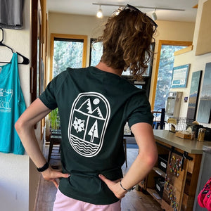 Man showing the back of t-shirt, which has a modern design with a mountain, ✿, tree, and river in it 