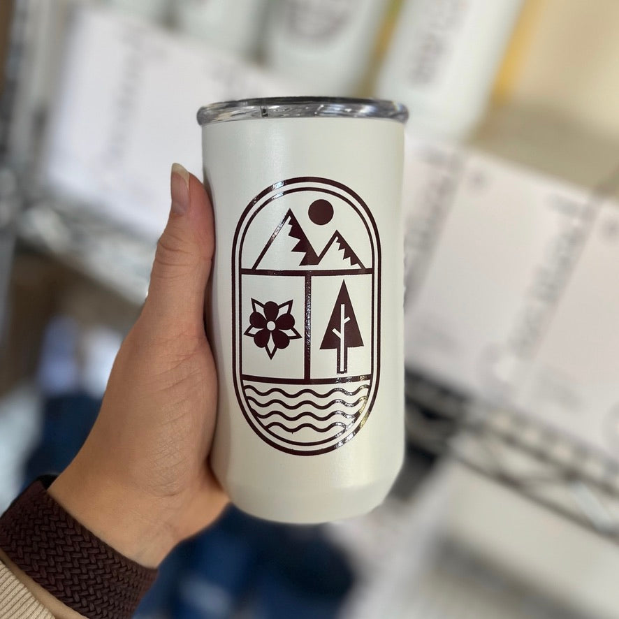 A person's hand holding out a white mug with a modern mountain, tree, and water design