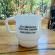 Load image into Gallery viewer, A white ceramic coffee mug with black printed text reading '470 E Elkhorn Ave, Estes Park, Colorado kindcoffee.com', sitting on a wooden table in front of a window with a forest view.
