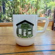 Load image into Gallery viewer, A white ceramic coffee mug with a handle, featuring a printed illustration of a coffee shop named 'KIND COFFEE' in green and black, sitting on a wooden table with a blurred outdoor forest background.
