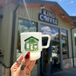 Load image into Gallery viewer, A hand with red nail polish holds a white ceramic coffee mug featuring a green and black graphic of a coffee shop building with the text 'KIND COFFEE'. The background shows the exterior of a building with a matching 'KIND COFFEE' sign, in a sunny outdoor setting.
