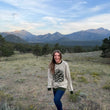 Load image into Gallery viewer, A woman stands in a grassy mountain meadow with pine trees and mountains in the background, wearing a cream knit sweater with a large pinecone design and dark green ribbed trim, paired with dark blue jeans.
