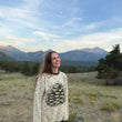 Load image into Gallery viewer, A woman stands outdoors in a grassy field with mountains and trees in the background, wearing a cream long-sleeve sweater with a pinecone graphic and small dark heart-like dots.
