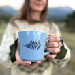 Load image into Gallery viewer, A person outdoors holds up a light blue ceramic mug with a black fern leaf design printed on the side. The background is a soft-focus natural landscape with mountains and grass.
