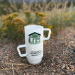 Load image into Gallery viewer, Two stacked white ceramic mugs with handles, printed with a green and black Kindo Coffee logo and address, photographed outdoors on gravel with blurred wildflowers in the background.
