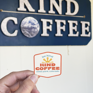 Be Kind sticker held in front of a Kind Coffee wall sign.