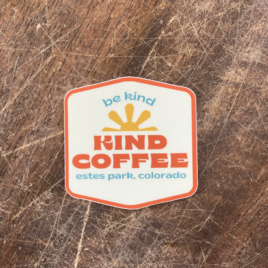 Be Kind sticker on a wooden table. Sticker has a white background and orange border. Text reads "be kind - KIND COFFEE - estes park, colorado"